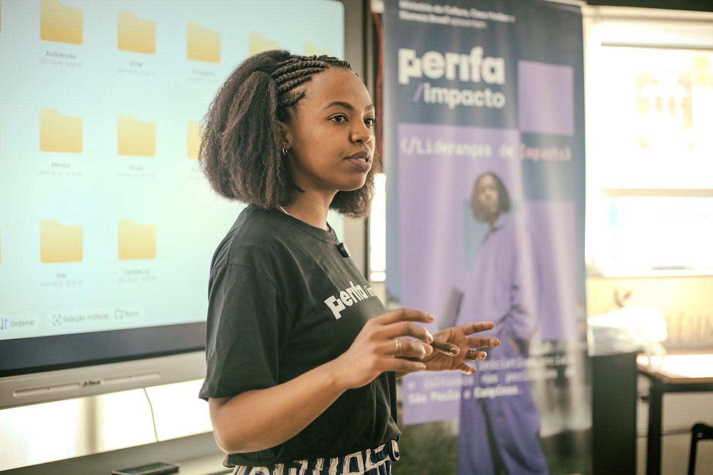 Joven negra con trenzas, con camiseta de Perifa Impacto, presentando frente a una pantalla en una formación de Casa Hacker