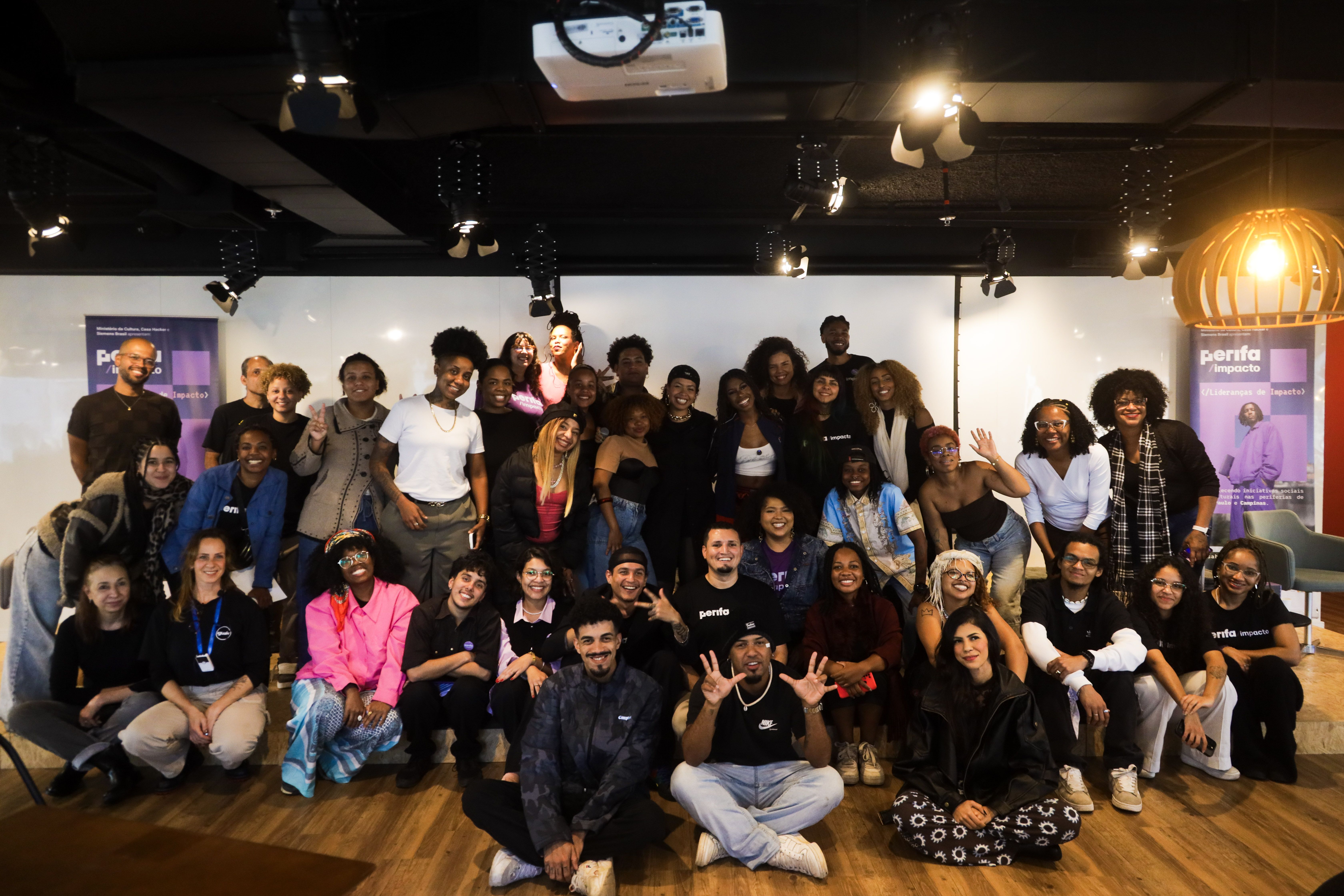 Cerca de cinquenta lideranças de impacto reunidas para a foto de grupo em um salão, durante encontro do PerifaImpacto — ao fundo, roll-ups do programa com as marcas do Ministério da Cultura, da Casa Hacker e da Siemens Brasil