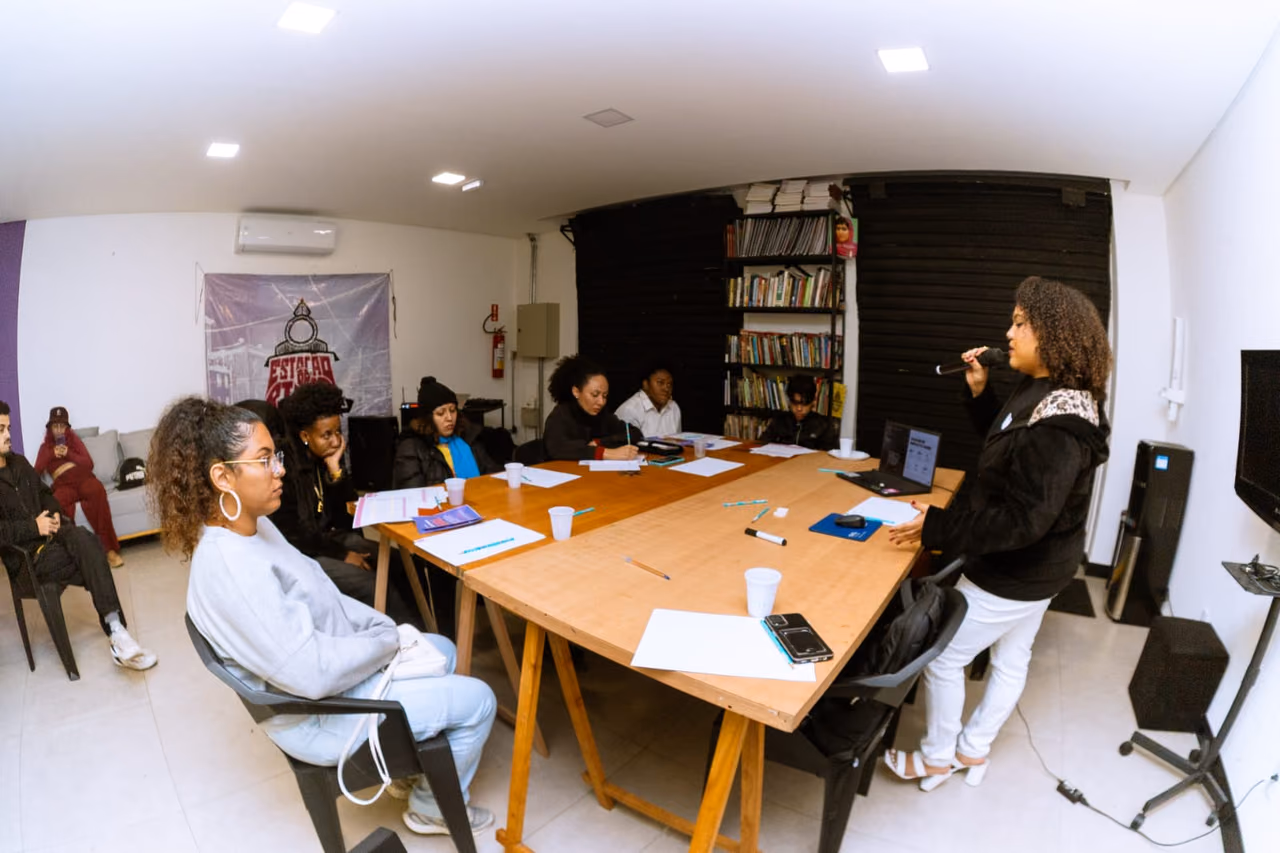 Participantes de uma atividade dos Hubs de Cidadania Ativa reunidos em torno de uma mesa enquanto uma facilitadora conduz a atividade. O grupo está em um espaço de formação, com materiais de trabalho, notebooks e uma biblioteca ao fundo.