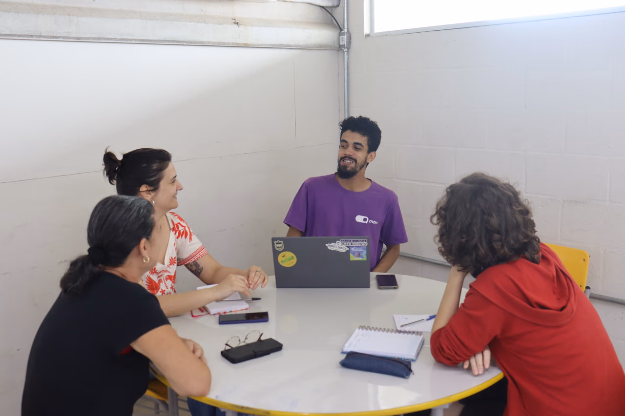 Four people talking around a round table with a laptop, notebooks, and cell phones in a room with white walls.
