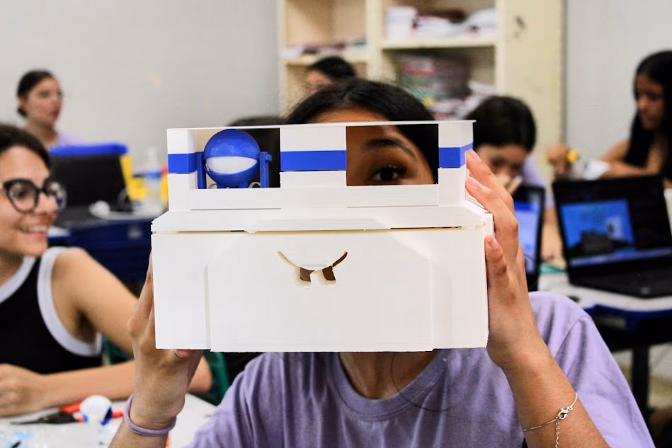 A student holds a white prototype with blue details in front of their face, built during a Hackerclubes activity. In the background, other students work on computers and experimentation materials.