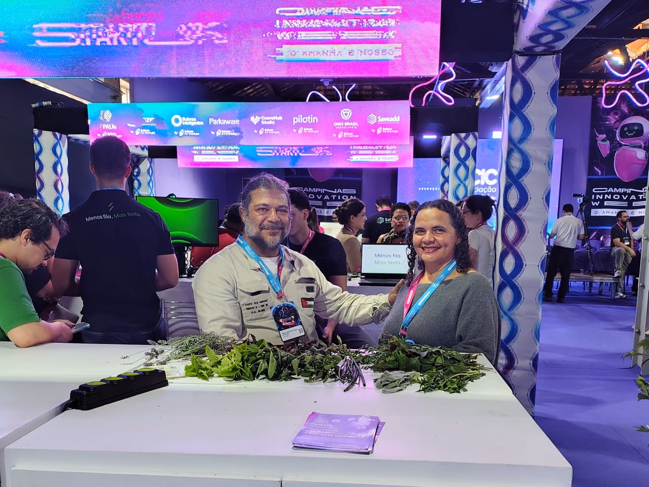 Fátima and Daniel, from Horta do Itajaí, pose side by side in a space at Campinas Innovation Week 2026. In front of them, there is a table with different types of leaves and herbs. In the background, booths, screens, and colorful elements of the event appear.
