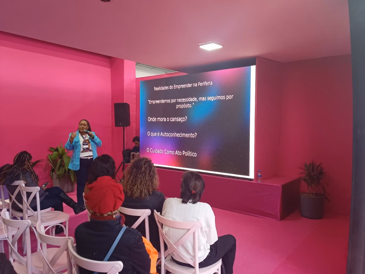 Karla Marques speaks into a microphone during a workshop, in front of a seated audience. In the background, a screen displays the theme “Realities of Entrepreneurship in the Periphery” and topics on purpose, self-care, and self-knowledge.