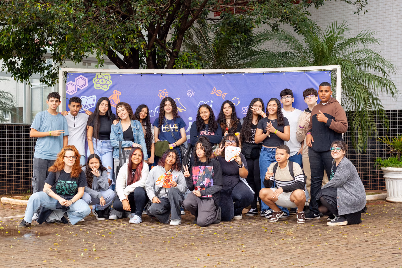 Grupo de estudantes e integrantes do Hackerclubes posa para uma foto em frente a um painel decorado com elementos relacionados à ciência e à tecnologia. Os participantes estão reunidos em diferentes posições, alguns em pé e outros sentados, durante a II Feira de Ciências InterCOCEN.