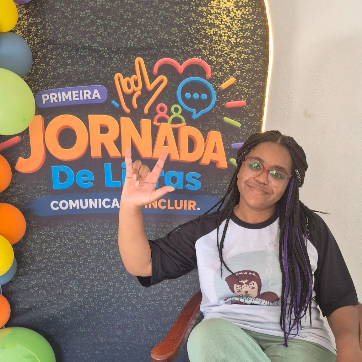Helen, assistente de comunicação do Hub Quebrada em Movimento, está sentada diante de um painel da Primeira Jornada de Libras e faz um sinal em Libras com a mão. Balões coloridos decoram o espaço ao redor do painel.