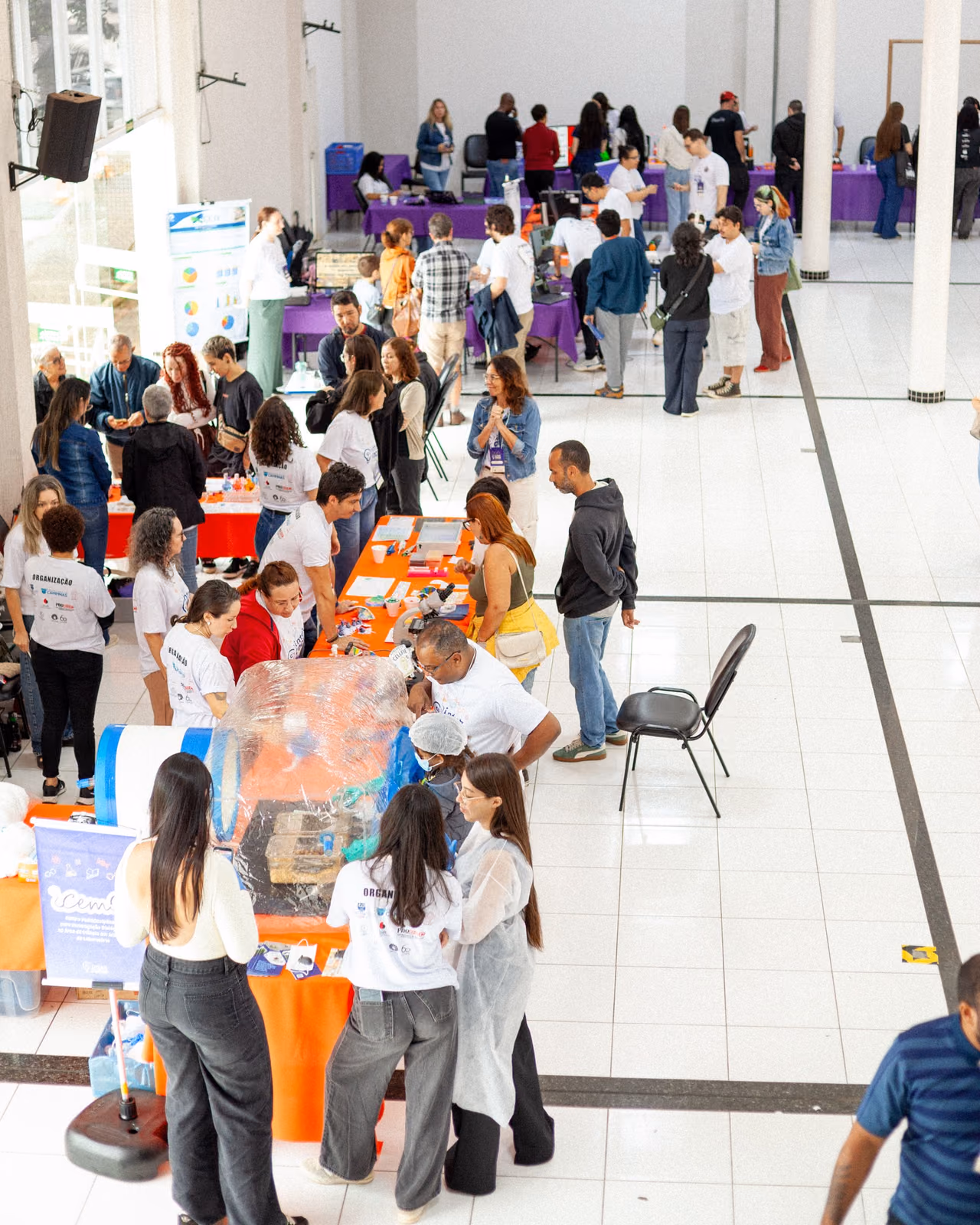 Vista geral da II Feira de Ciências InterCOCEN, realizada em um amplo espaço com diversos estandes e mesas de exposição. Participantes circulam pelo local, conversam com os expositores e conhecem projetos e experimentos apresentados.