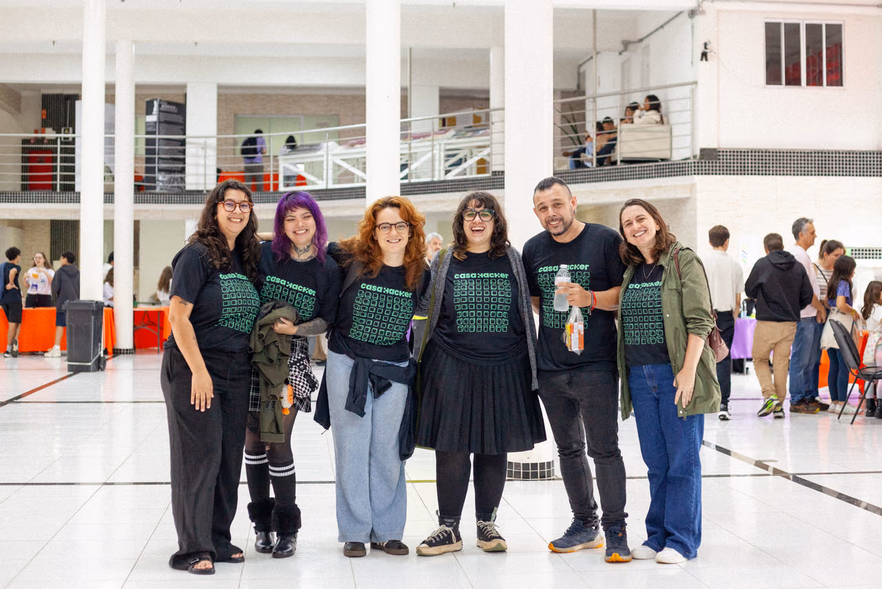 Seis integrantes da Casa Hacker posam juntos no espaço da II Feira de Ciências InterCOCEN. Eles usam camisetas pretas com a identidade visual da organização e estão em um salão com estandes e participantes do evento ao fundo.
