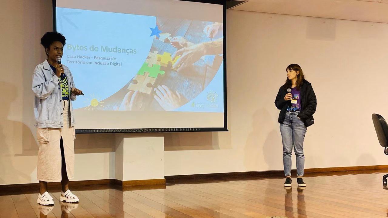 A fotografia registra um momento de apresentação no palco de um auditório, onde duas representantes da Casa Hacker compartilham os resultados da pesquisa “Bytes de Mudanças”. À esquerda, uma apresentadora segura o microfone e gesticula durante a explanação, vestindo jaqueta jeans clara sobre camiseta estampada e calça bege, com calçado branco estilo crocs. À direita, sua colega acompanha a apresentação com microfone em mãos, vestindo jaqueta preta, camiseta roxa com identificação do evento, calça jeans e tênis de plataforma preto.
O slide projetado exibe o título “Bytes de Mudanças” com o subtítulo “Casa Hacker - Pesquisa de Território em Inclusão Digital”, acompanhado de uma imagem ilustrativa de mãos montando peças de quebra-cabeça em tons pastéis (azul, verde, amarelo e bege), simbolizando a construção colaborativa e a conexão entre diferentes atores sociais. O ambiente é um auditório formal com piso de madeira, paredes claras e estrutura adequada para eventos de grande porte.