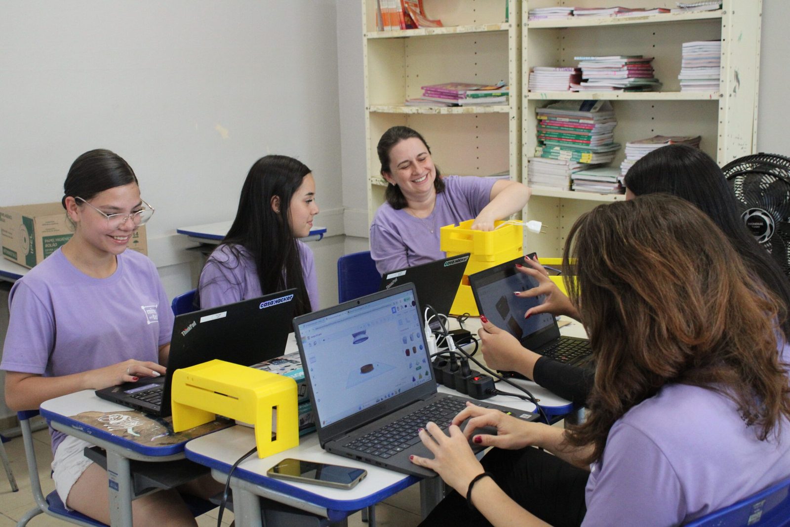 grupo de quatro alunas sentadas em sala de aula com seus computadores. Na mesa na frente delas, além dos computadores vemos peças de impressão 3D amarelas. Entre as alunas está sentada a Professora do grupo. Todas estão sorrindo.