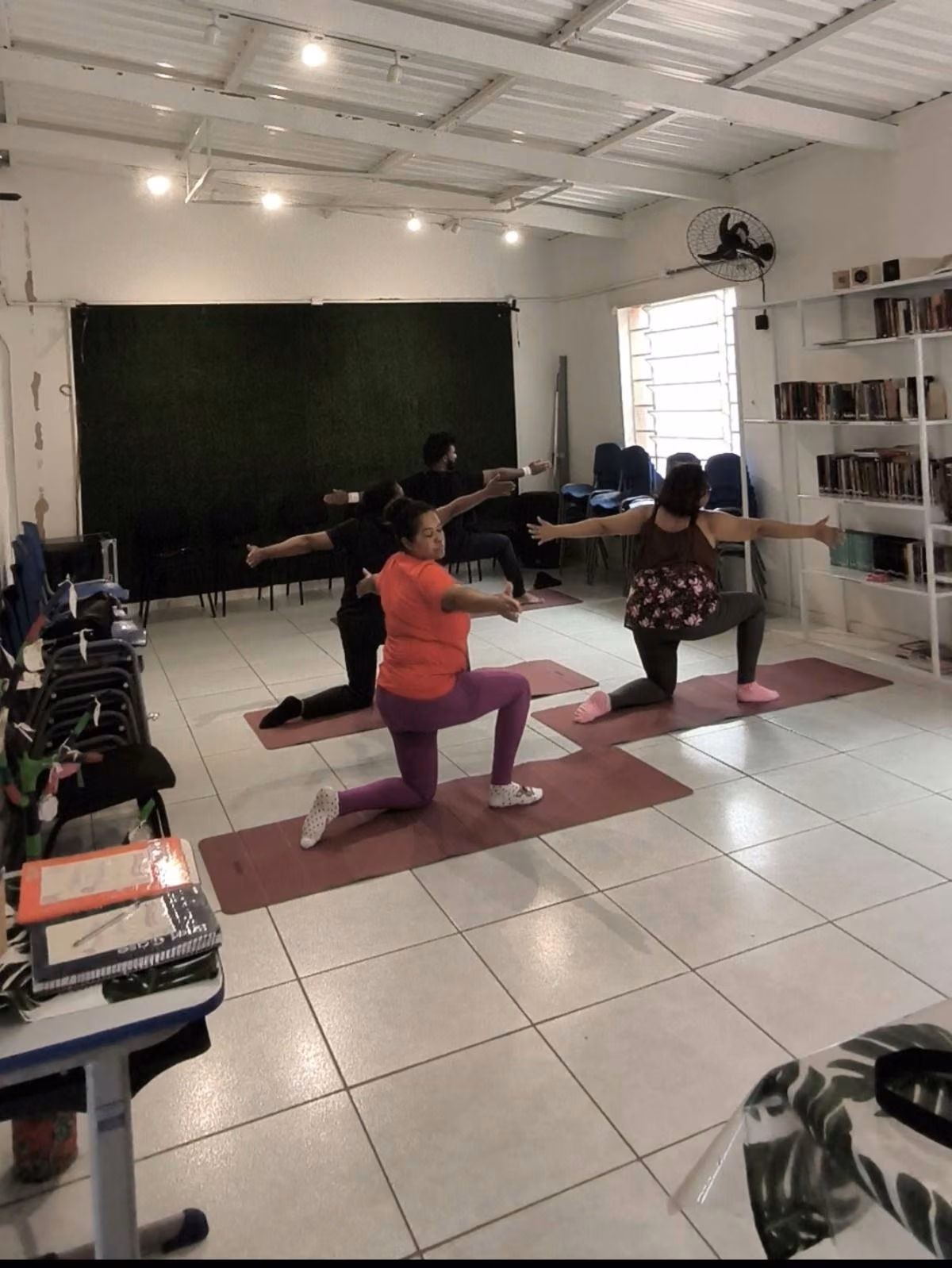 Três pessoas fazem uma postura de ioga sobre tapetes, numa sala com estante de livros ao fundo.