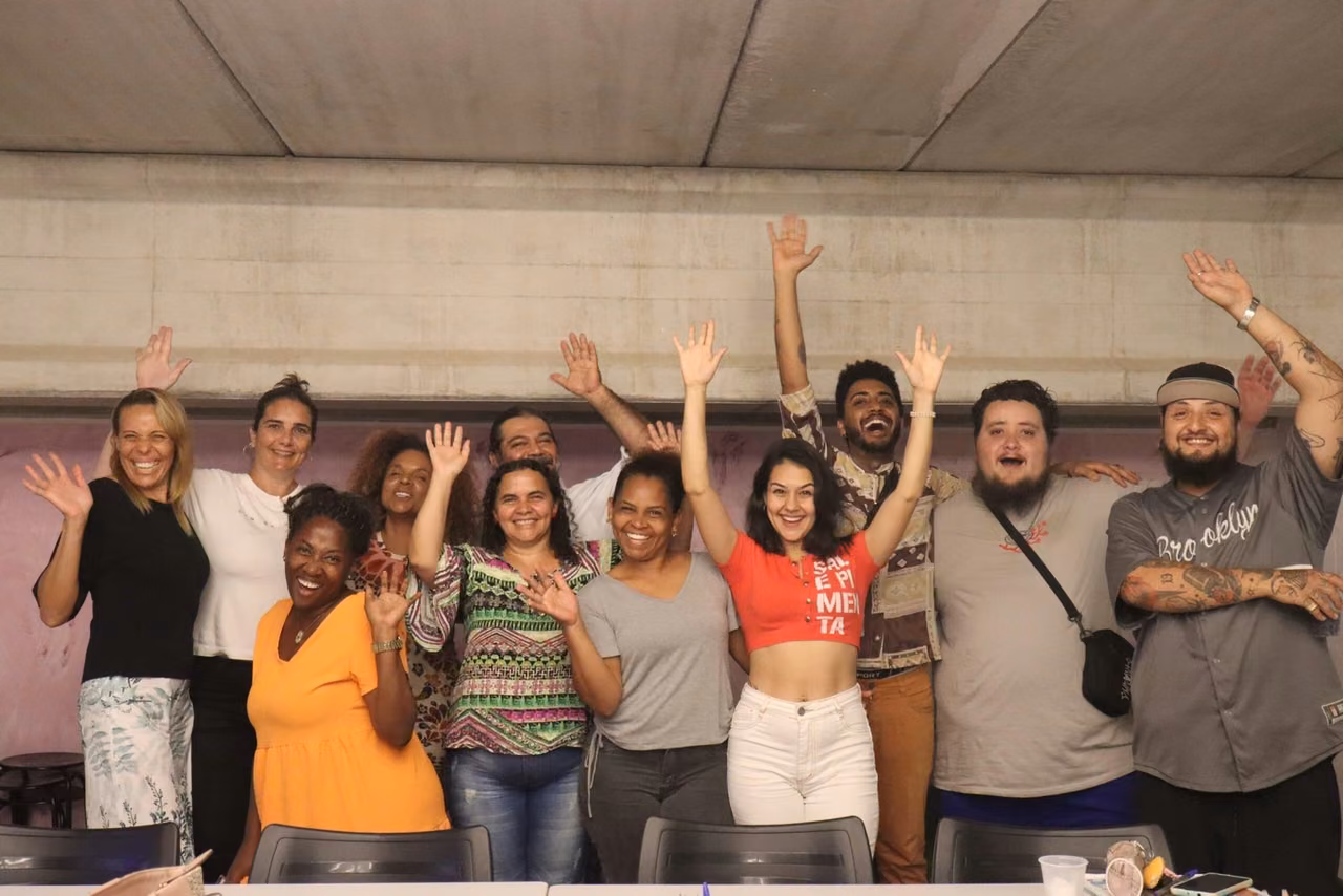 Doze pessoas de braços erguidos e sorrindo, reunidas em volta de uma mesa num espaço comunitário.