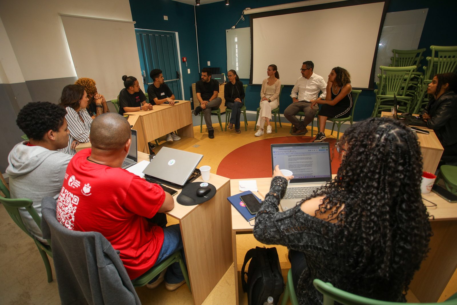 Roda de conversa em sala do Hub: participantes com notebooks sentados em círculo de mesas de madeira acompanham a fala de convidados.