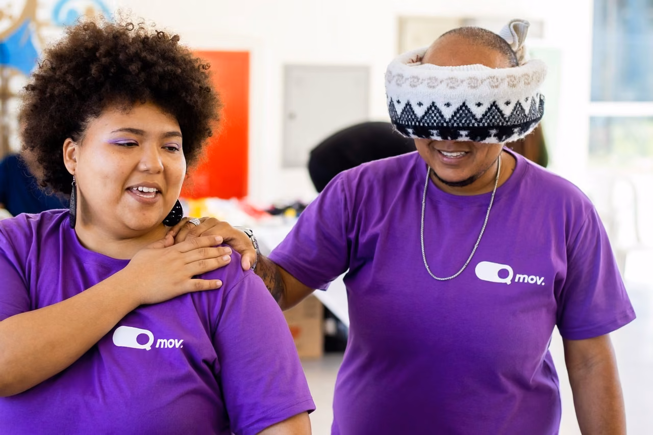Dois participantes de uma atividade interativa vestem camisetas roxas com o logotipo 'Q Mov.'. A pessoa à esquerda, uma mulher com cabelo crespo volumoso e maquiagem roxa, sorri enquanto guia a pessoa à direita, que usa um gorro puxado sobre os olhos como uma venda. Ela apoia a mão no ombro do colega, que sorri enquanto segura suavemente seu ombro. O ambiente parece ser um espaço interno iluminado, possivelmente um evento ou oficina.
