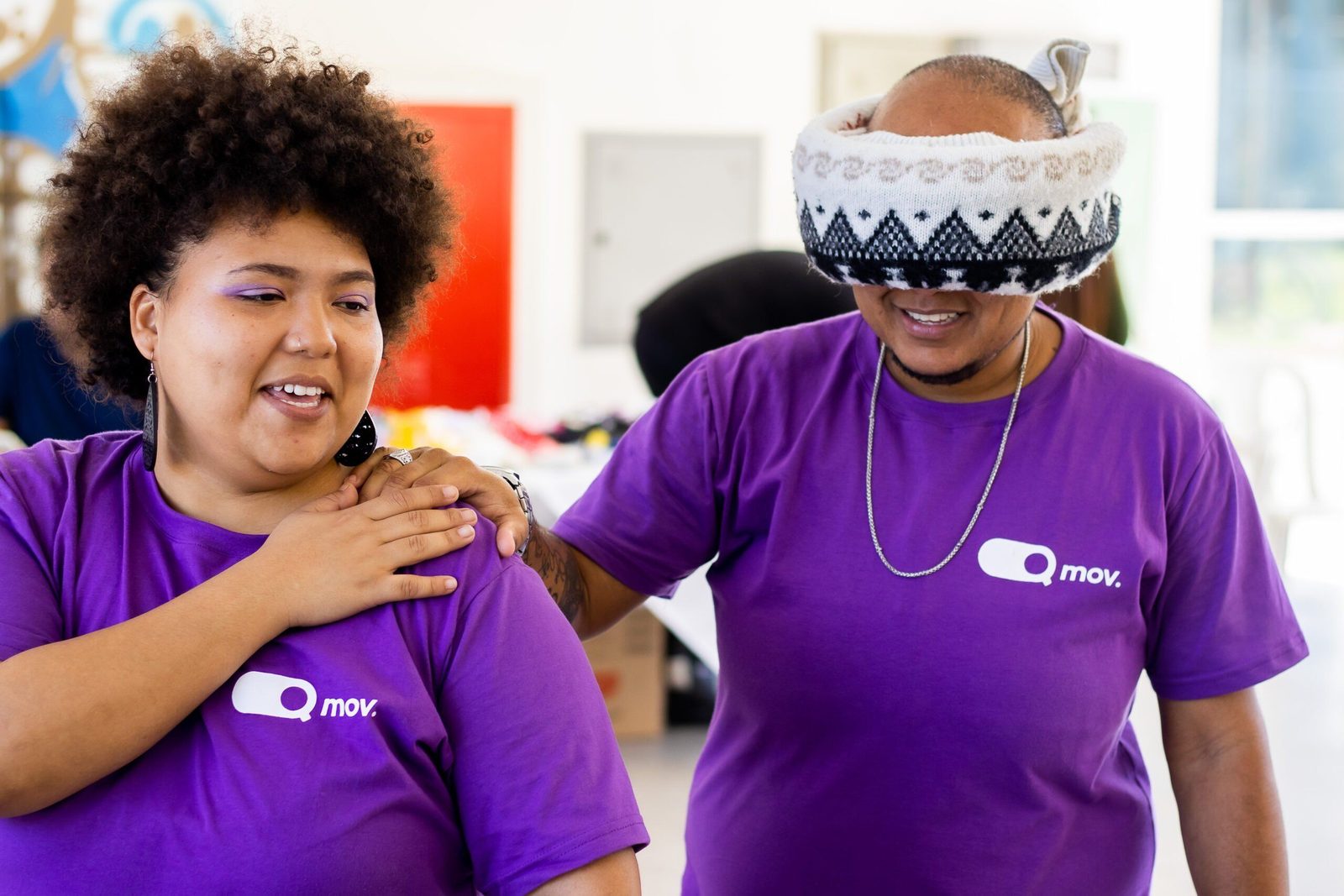 Dois participantes de uma atividade interativa vestem camisetas roxas com o logotipo 'Q Mov.'. A pessoa à esquerda, uma mulher com cabelo crespo volumoso e maquiagem roxa, sorri enquanto guia a pessoa à direita, que usa um gorro puxado sobre os olhos como uma venda. Ela apoia a mão no ombro do colega, que sorri enquanto segura suavemente seu ombro. O ambiente parece ser um espaço interno iluminado, possivelmente um evento ou oficina.