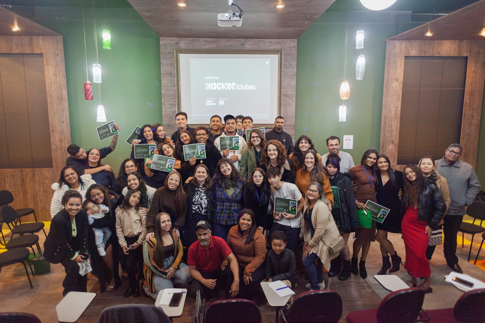 Estudantes e familiares reunidos no auditório seguram certificados diante do telão com a vinheta da formatura do Hackerclubes.