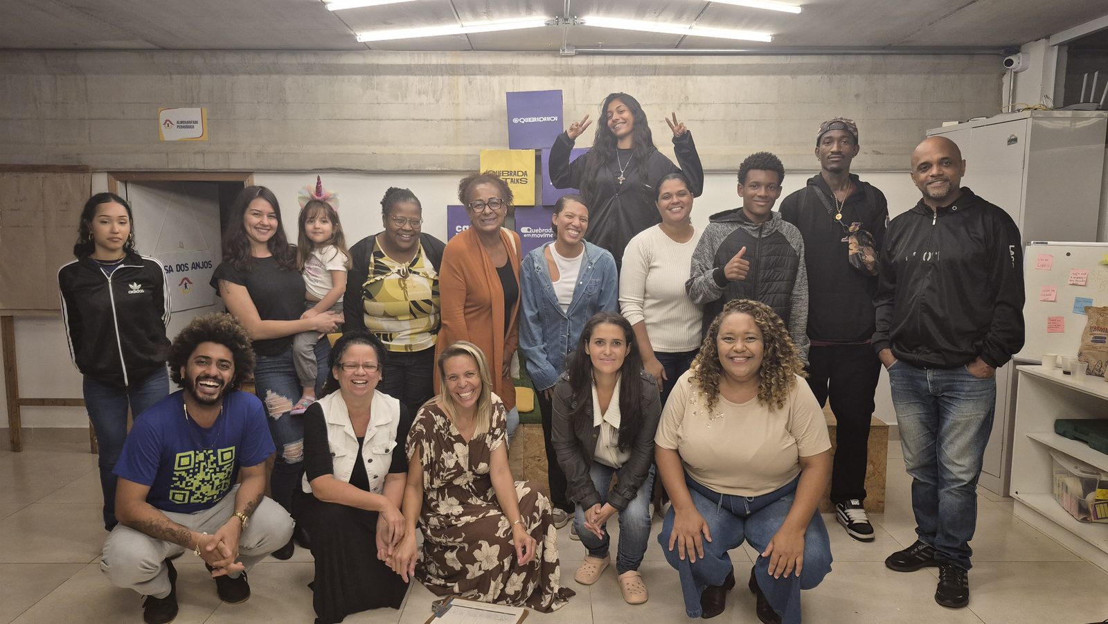 Moradores e lideranças de várias gerações posam juntos no espaço do Hub Quebrada em Movimento, com caixas com o logotipo do programa ao fundo.