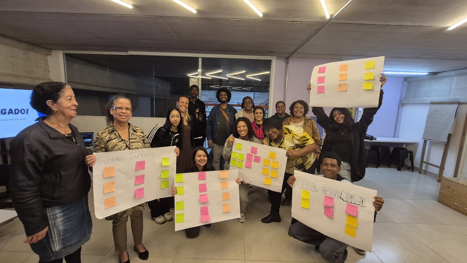 Participantes da jornada formativa exibem cartazes com as colunas fazer, fazendo e feito, preenchidas com post-its coloridos.