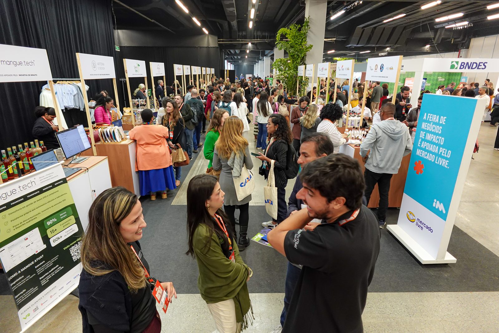 Pavilhão lotado do evento Impacta Mais, com estandes de negócios de impacto, totens do Mercado Livre e do BNDES e grupos de pessoas com crachás conversando.