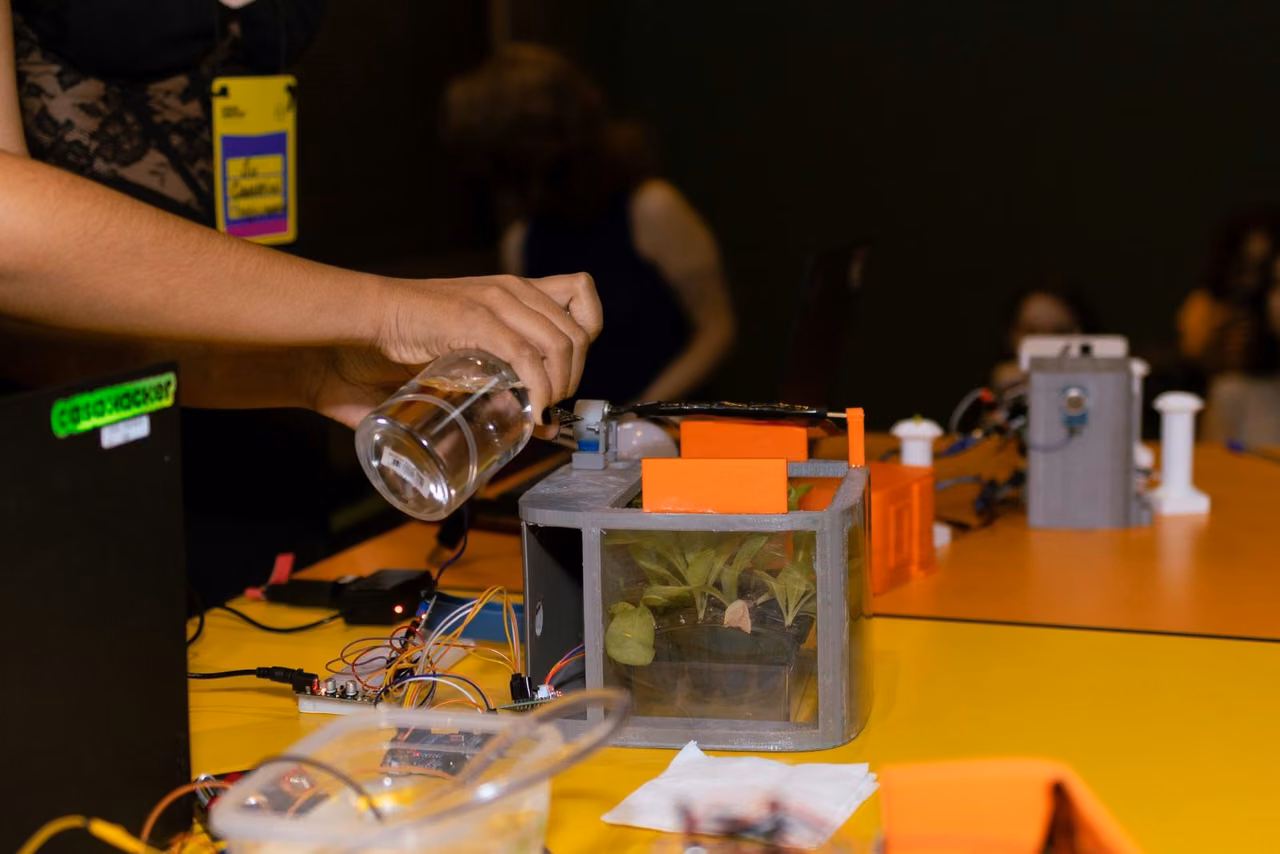 Mão despeja água de um béquer num protótipo de horta com plantas, peças impressas em 3D e fios numa protoboard.