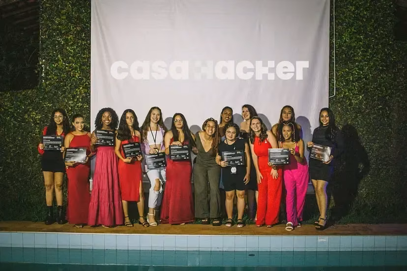Doze meninas enfileiradas seguram certificados diante de um painel com a marca Casa Hacker.