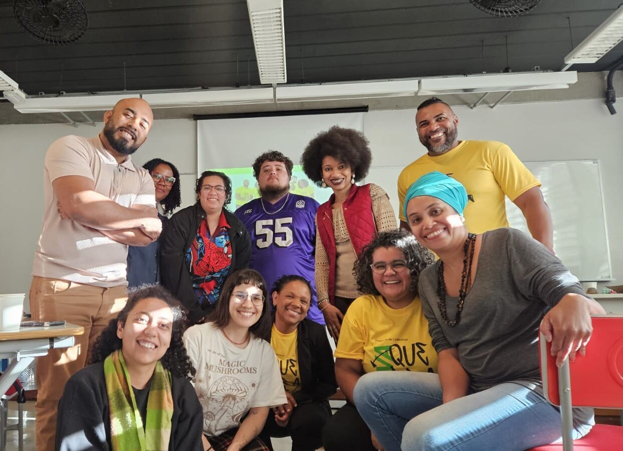 Grupo de lideranças do Inova Quebrada posa sorrindo em sala de aula, em pé e sentadas diante de um telão de projeção.