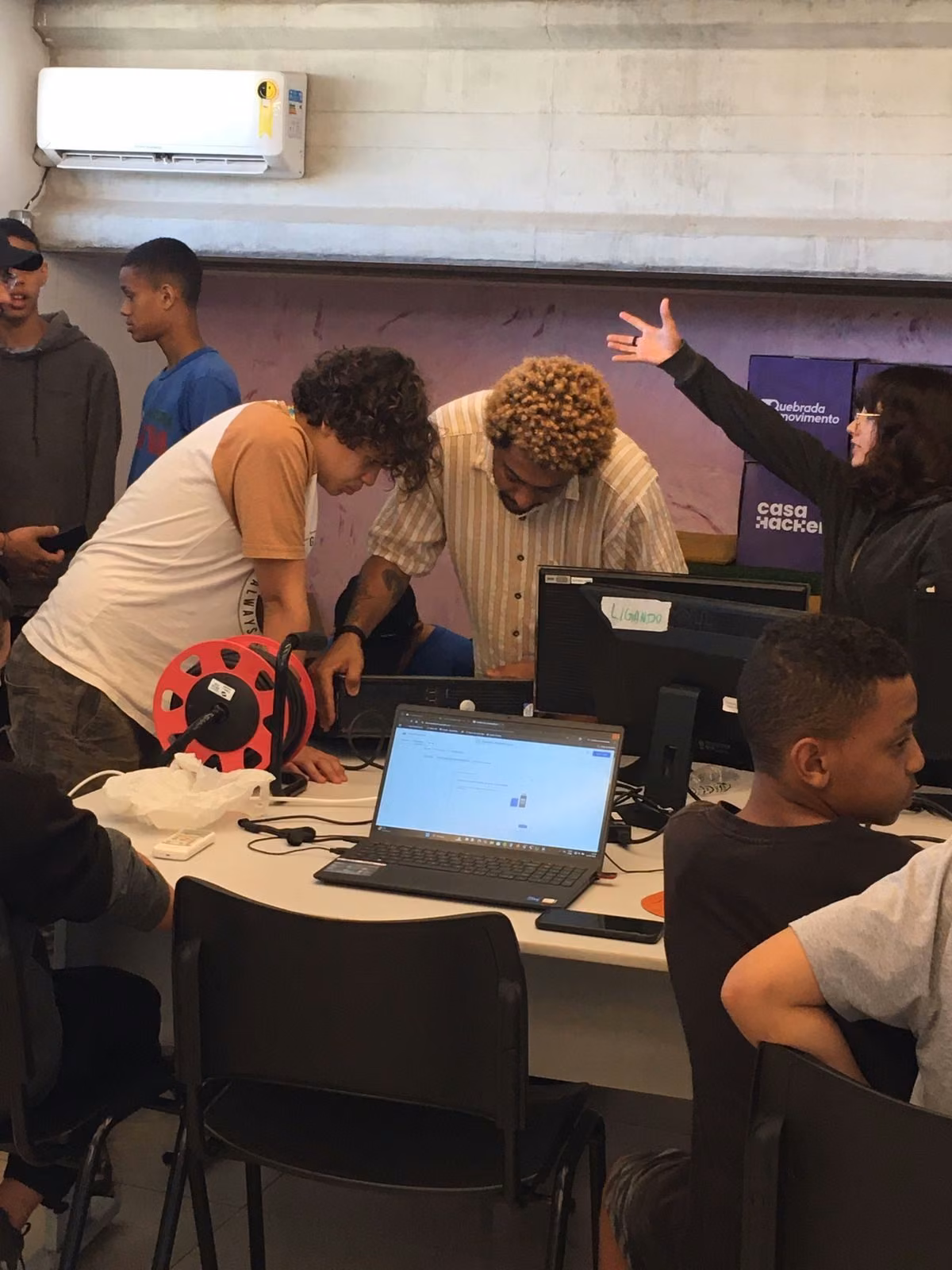 Jovens em volta de uma bancada com notebook, monitor e bobina de filamento na oficina do Hub.