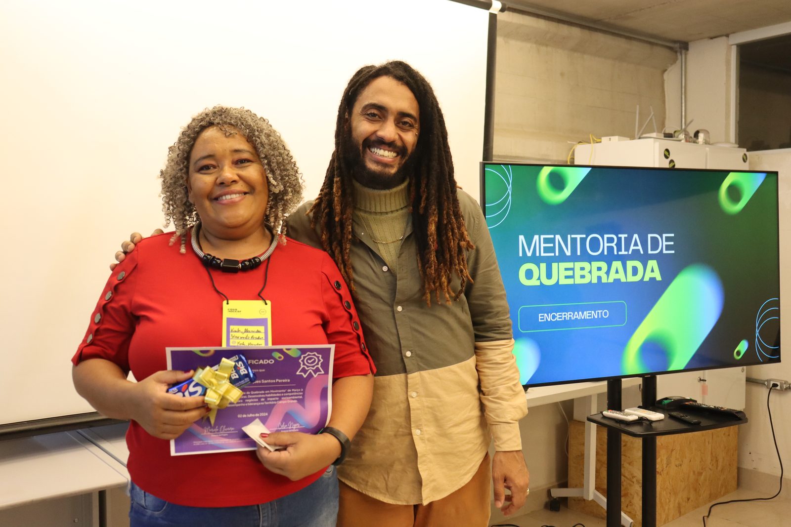 Mulher sorridente segura certificado e chocolates ao lado de homem de dreadlocks; ao fundo, TV exibe o slide Mentoria de Quebrada, encerramento.