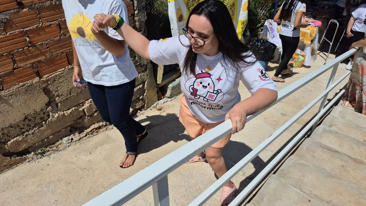 Mulher de óculos sobe a rampa de concreto recém-construída apoiada no corrimão metálico branco, amparada por outra participante, na sede da ONG Gabriel.