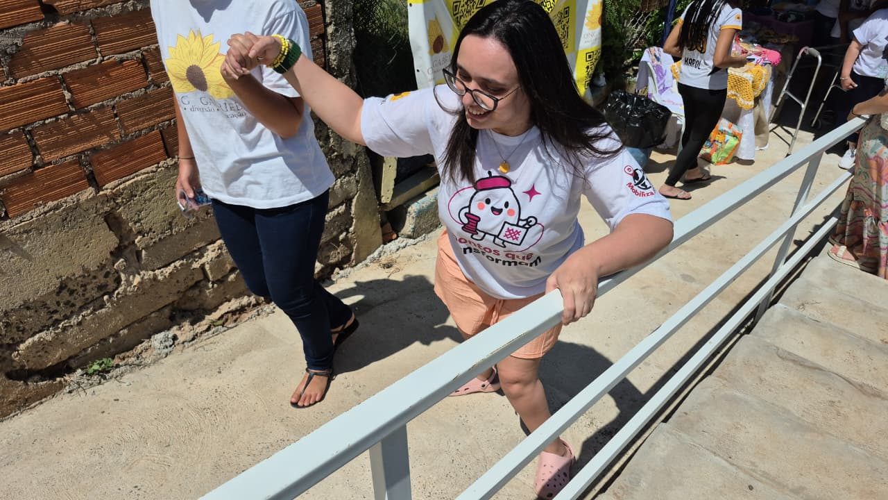 Mulher de óculos sobe a rampa de concreto recém-construída apoiada no corrimão metálico branco, amparada por outra participante, na sede da ONG Gabriel.