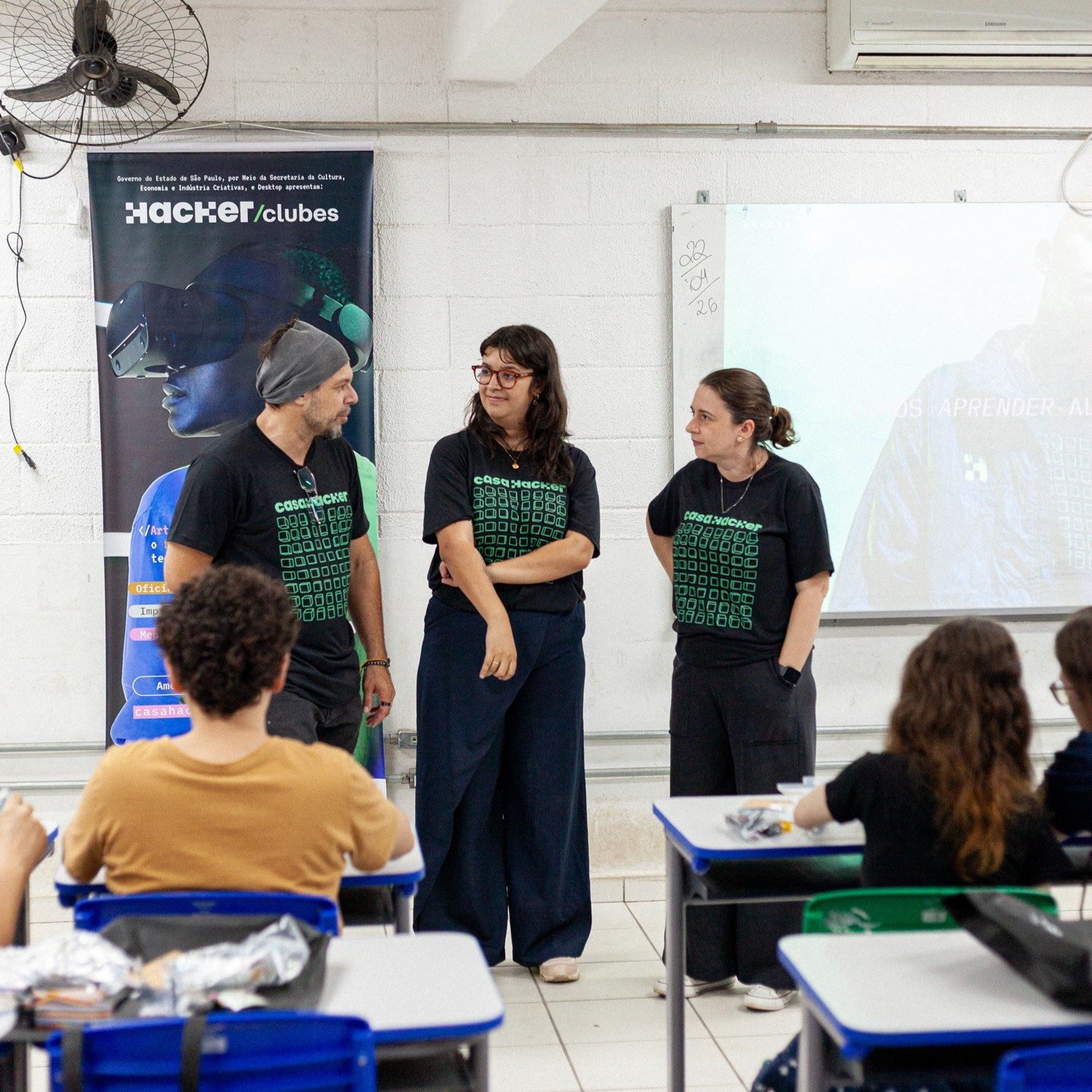 Três educadores com camisetas pretas da Casa Hacker conversam em pé diante de uma turma sentada em carteiras escolares, ao lado de um banner do Hackerclubes.