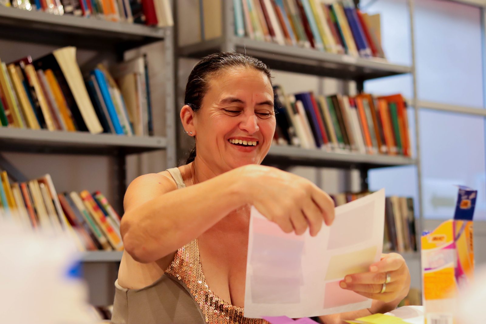 Mulher sorrindo enquanto lê um papel durante uma atividade em grupo, sentada em um espaço com estantes repletas de livros ao fundo.