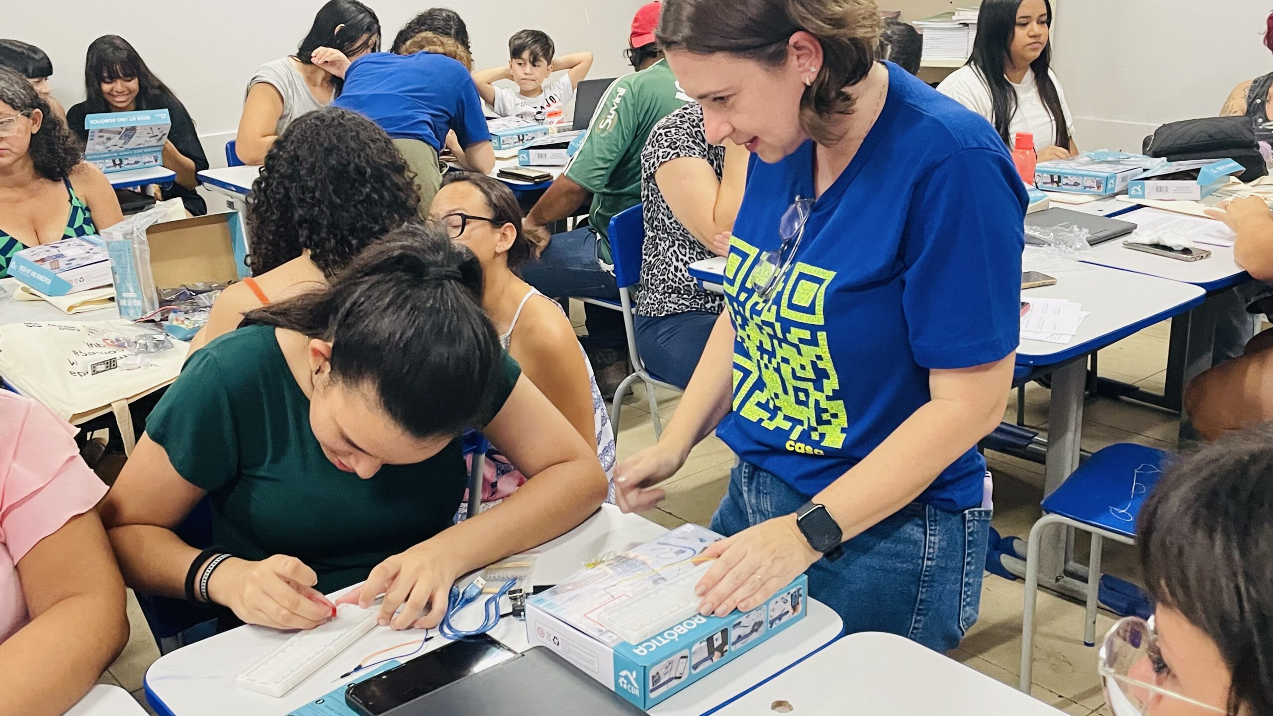 Educadora em camiseta azul com logo Hacker Clubes orienta adolescente em atividade hands-on com kit educativo. Sala com múltiplos participantes, mesas colaborativas com materiais de aprendizagem. Ambiente acolhedor onde aprendizagem acontece através de interação genuína e exploração prática.