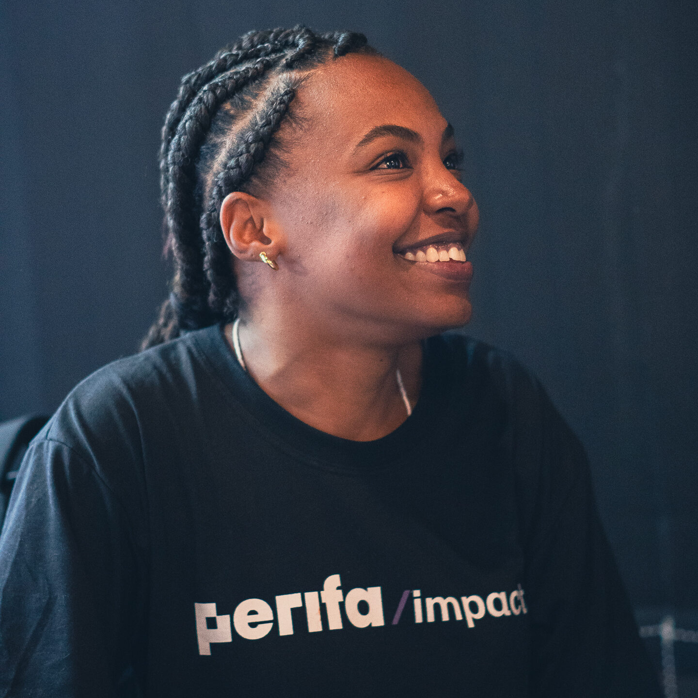 Mulher negra jovem, de perfil, sorri enquanto olha para o alto, à direita da imagem. Ela usa o cabelo trançado para trás, brincos dourados pequenos e uma camiseta preta com a inscrição “perifa/impacto” em letras brancas. O fundo é escuro e desfocado, destacando seu rosto iluminado e sua expressão de alegria e atenção.