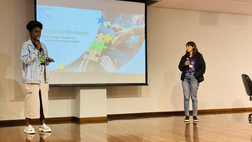 A fotografia registra um momento de apresentação no palco de um auditório, onde duas representantes da Casa Hacker compartilham os resultados da pesquisa "Bytes de Mudanças". À esquerda, uma apresentadora segura o microfone e gesticula durante a explanação, vestindo jaqueta jeans clara sobre camiseta estampada e calça bege, com calçado branco estilo crocs. À direita, sua colega acompanha a apresentação com microfone em mãos, vestindo jaqueta preta, camiseta roxa com identificação do evento, calça jeans e tênis de plataforma preto.
O slide projetado exibe o título "Bytes de Mudanças" com o subtítulo "Casa Hacker - Pesquisa de Território em Inclusão Digital", acompanhado de uma imagem ilustrativa de mãos montando peças de quebra-cabeça em tons pastéis (azul, verde, amarelo e bege), simbolizando a construção colaborativa e a conexão entre diferentes atores sociais. O ambiente é um auditório formal com piso de madeira, paredes claras e estrutura adequada para eventos de grande porte.