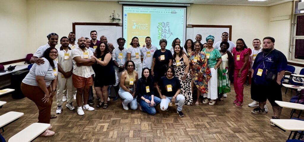 Registro coletivo de um encontro formativo realizado em uma sala universitária. A imagem reúne dezenas de participantes posicionados para uma foto em grupo, em um ambiente marcado pela diversidade racial, geracional e estética. Ao fundo, um projetor exibe a identidade visual do evento, reforçando o caráter colaborativo e comunitário da atividade. A composição transmite acolhimento, troca de saberes e construção coletiva.