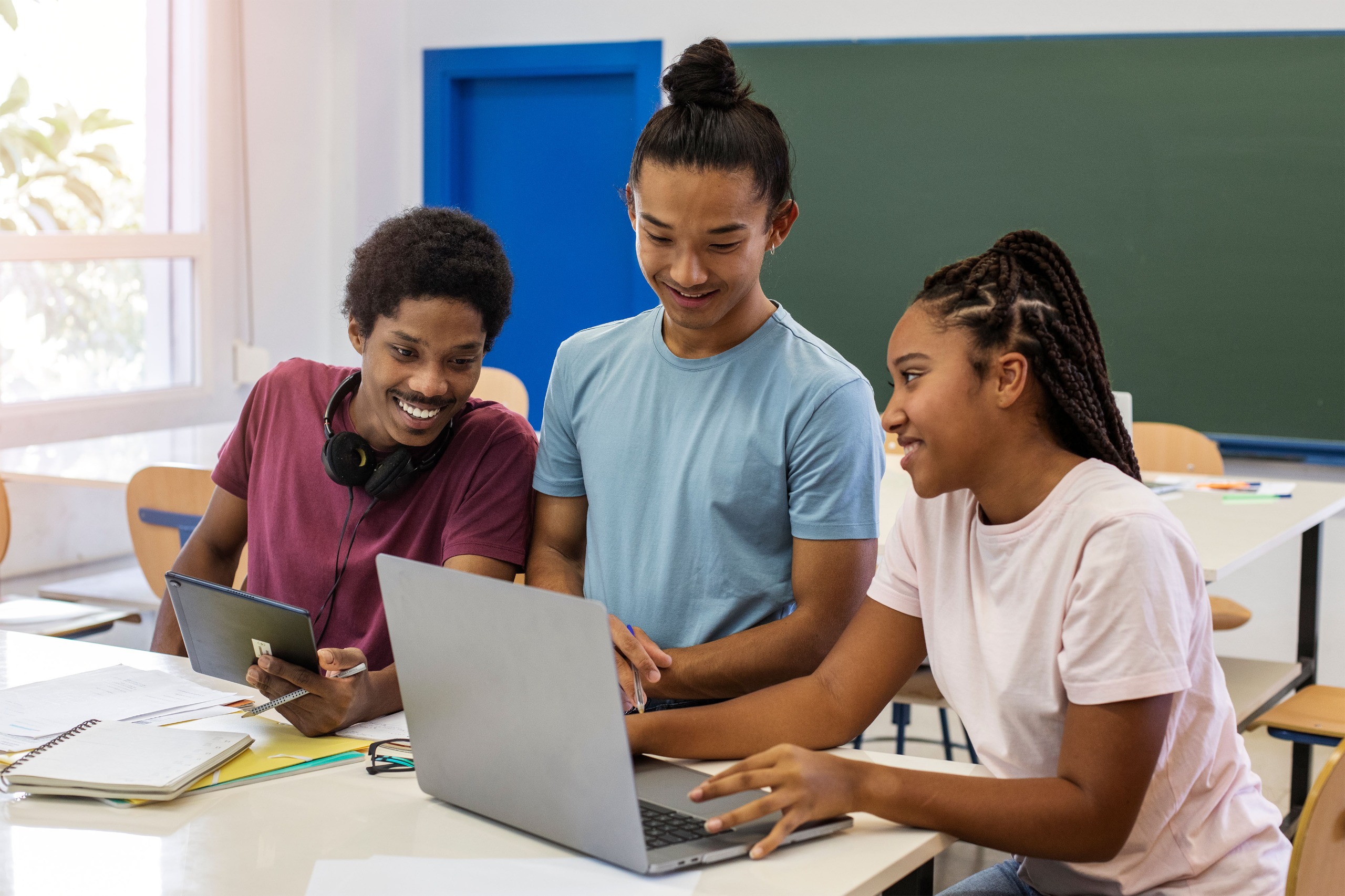 Três jovens estudantes em uma sala de aula colaboram em frente a um notebook, enquanto um deles segura um tablet e todos observam a tela com atenção e entusiasmo.