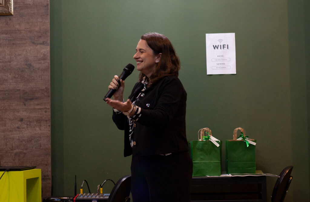 Aliene Villaça, em pé, falando ao microfone. Ela sorri enquanto fala.